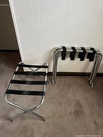 Two silver metal luggage racks with black fabric straps, one set up and one folded against a white wall and beige carpet.