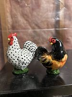 Photo of two ceramic chicken-shaped salt and pepper shakers, one white with black spots and the other black with orange and yellow coloring, on a black surface.