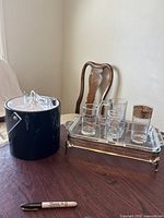 Black cylindrical ice bucket with clear plastic lid next to rectangular glass tray with gold finish frame holding six assorted drink and shot glasses on a wooden table.
