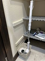 Electric tile cleaning wand stored upright in a beige fabric bin on the floor, with shelves in the background showing additional cleaning supplies and accessories.