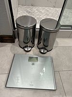 Photo showing two stainless steel Simple Human pedal trash cans and one Taylor digital bathroom scale with glass platform in front of a tiled shower