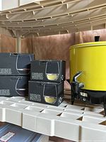 Yellow Chilton beverage dispenser standing next to stacked box sets of Riedel drinking glasses on a white plastic shelf.