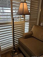 Floor lamp shown near a window with white blinds, brown fabric shade illuminated, metal pole and base visible.