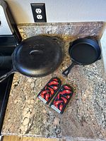 Large and small cast iron skillets displayed on countertop with large skillet lid and two red chili pepper patterned hot handle covers