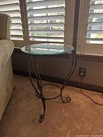 Circular side table with removable clear glass top and metal base, shown from side near window and armchair.
