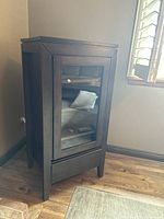 Dark brown wooden media cabinet pictured showing glass front door and drawer beneath, standing on hardwood floor near window.