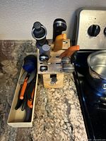 Wooden knife block with six knives of varying handle colors and a blade sharpener on the block, along with an organizing tray holding additional cutting utensils with orange and black handles