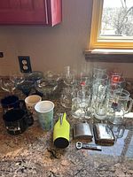 Photo showing assorted drinking glasses including pint glasses, beer mugs, shot glasses along with four ceramic mugs and a lime green bottle coozie with two metal flasks and a bottle opener on kitchen counter.
