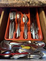 Open kitchen drawer showing orange plastic utensil organizer filled with assorted forks, spoons, and knives. Additional serving spoons and miscellaneous utensils are placed in front of the organizer.