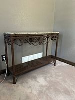 Side angle view of the entryway table showing the brown metal frame with decorative scrollwork and the granite top, placed against a beige wall on carpeted floor.