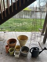 Photo showing multiple flower pots of ceramic and plastic in various sizes and colors, along with three metal tomato tresses and part of a black watering can on a stone patio under a staircase.