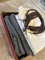 Folded gray and white curtain panels in a box next to five brown braided rope curtain pulls with loop ends and wooden beads.