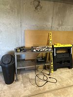 Photo showing metal work table, black trash can, yellow 48-inch level, and black plastic work horses.