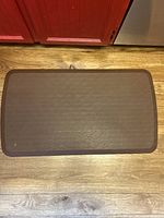 Top down view of rectangular brown kitchen floor mat placed on wood flooring, showing texture and condition.