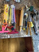Full overview of kitchen utensils, gadgets including plastic wrap box, reusable straws in pack, rolling pin, chip clips, baster, measuring cups, metal spoons, and grilling utensils arranged on countertop.