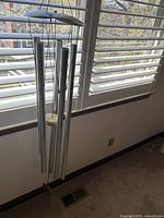 Front view of large silver wind chime with six metal tubes and wood accent top, hanging indoors in front of white slatted window blinds