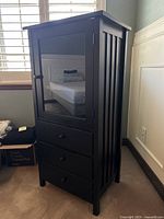 Front-side angle view of dark wood media cabinet showing glass door with two inner shelves and three lower drawers.