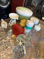 Photo showing all miscellaneous kitchen items together on counter including salad spinner, honey pot, manual food processors, scale, measuring cups, sifter, sugar dispenser, butter tray.