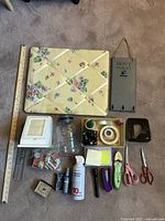 Photo showing floral fabric-covered pushpin board with ribbons, 'Don't Forget' hanging organizer, and various office items including cutting tools, adhesives, and Pottery Barn bookend.