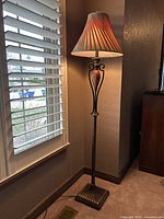Full view of the lamp showing base, column detail, and illuminated fabric shade lightly glowing, positioned near window with blinds.