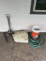 Photo showing the watering hose coiled next to two white 5-gallon buckets and a beige tarp on a patterned floor against a siding wall.