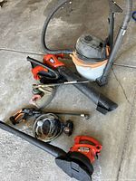 Photo showing multiple tools on concrete floor including two red and black Black & Decker leaf blowers, Ridgid shop vacuum with orange band and white body, Toro edger with metal blade, Worx Hydro Shot with hose coiled, and various hoses/attachments.