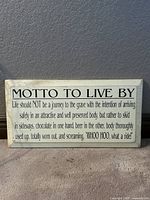 Front view of white wooden plaque with black text quote 'MOTTO TO LIVE BY' in bold and the rest of the humorous quote beneath.