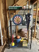 Wire shelving unit holds dart board, pennant, metal sign, cups, ball pump, baseball bat, basketball and baseballs, and other sports decor and gear.