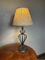 Side view of the table lamp turned on, showing the beige shade illuminated and the black metal wireframe base on a wooden surface.