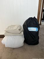 Photo of twin bedding items stacked including white quilted mattress protector pad, beige fitted sheet, beige flat sheet, and black AeroBed storage bag for inflatable mattress.