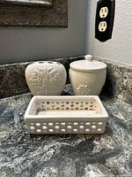 Set of three white ceramic bathroom organizers on a granite counter: toothbrush holder, container with lid, and small tray.