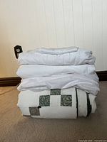 Stack of bedding items including the quilt with patchwork, white sheets, and pillowcase folded on carpet.