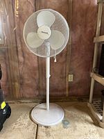 Front view of white Honeywell rotary fan showing three blades and pull cord control, standing on a concrete floor with an unfinished wall background.