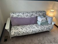 Futon with floral print mattress cover and bedding items stacked on one side including pillows, blankets, and sheets.