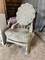 White wicker rocking chair with a round woven backrest showing a floral pattern, a hole cut in the seat, and a detached back cushion leaning against it. The chair appears used and located in basement.