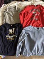Four shirts laid out on bed: grey quarter zip, red graphic 'Slap the Puck out of ALS', navy blue Milwaukee Brewers shirt, light blue textured shirt