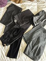Overview of mixed formal and informal women's bottoms in dark and neutral colors laid flat on a bed, featuring pants and skirts in various fabrics and styles.