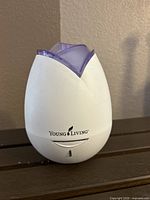 Front view of the Young Living essential oil diffuser, white body with purple top, showing logo and power indicator light.