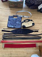 Photo showing two purses, one dark blue leather or faux leather with square cutout handle, one small beige and black purse, with multiple belts and two wide red scarf holders laid out on wooden floor.