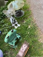 Outdoor photo showing the large metal flower ornament, green frog, and small brown bench decoration placed on grass near a paved path
