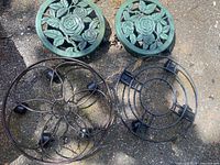 Overhead photo showing two green metal plant pot holders with decorative rose and leaf patterns, alongside two black plastic plant pot holders with simple open frames.