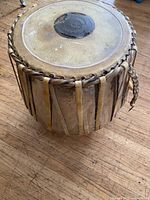Top view of antique metal drum showing natural hide head with central black patch and surrounding leather straps tied vertically down the sides.