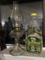 Clear glass oil lantern with tall chimney and curved base, placed next to a yellow capped bottle of Citrolite Outdoor Citronella Lamp Oil.
