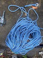 Coil of thick blue braided nylon rope with a metal eyelet secured by an orange tag at one end, lying on a rough outdoor surface with part of other items visible.