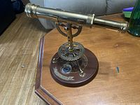 Angled side view showing polished brass telescope mounted on a circular wooden base with metal decor and a compass embedded.