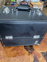 Closed black Caboodles cosmetic case showing leather texture, metal locks, handle and corner protectors.