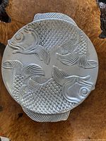 Cast metal oval serving plate with embossed fish design laid on textured wooden surface, shows detailed pattern and overall design.