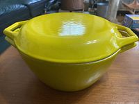 Yellow enamel coated cast iron Dutch oven with lid on, sitting on wooden table, showing both handles and the rounded shape.