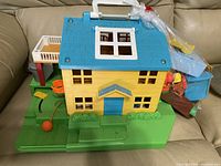 Closed view of yellow Fisher-Price house with blue roof, white handle, green base, and attached play elements.