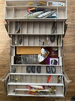 Top-down view of an open beige tackle box showing various fishing lures, weights, and accessories neatly arranged in compartments.